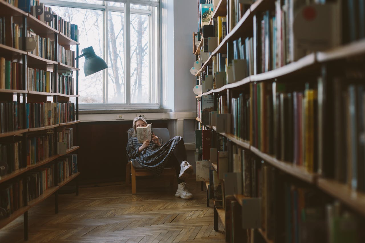 Woman enjoying a quiet read in a sunlit library corner with rows of bookshelves.